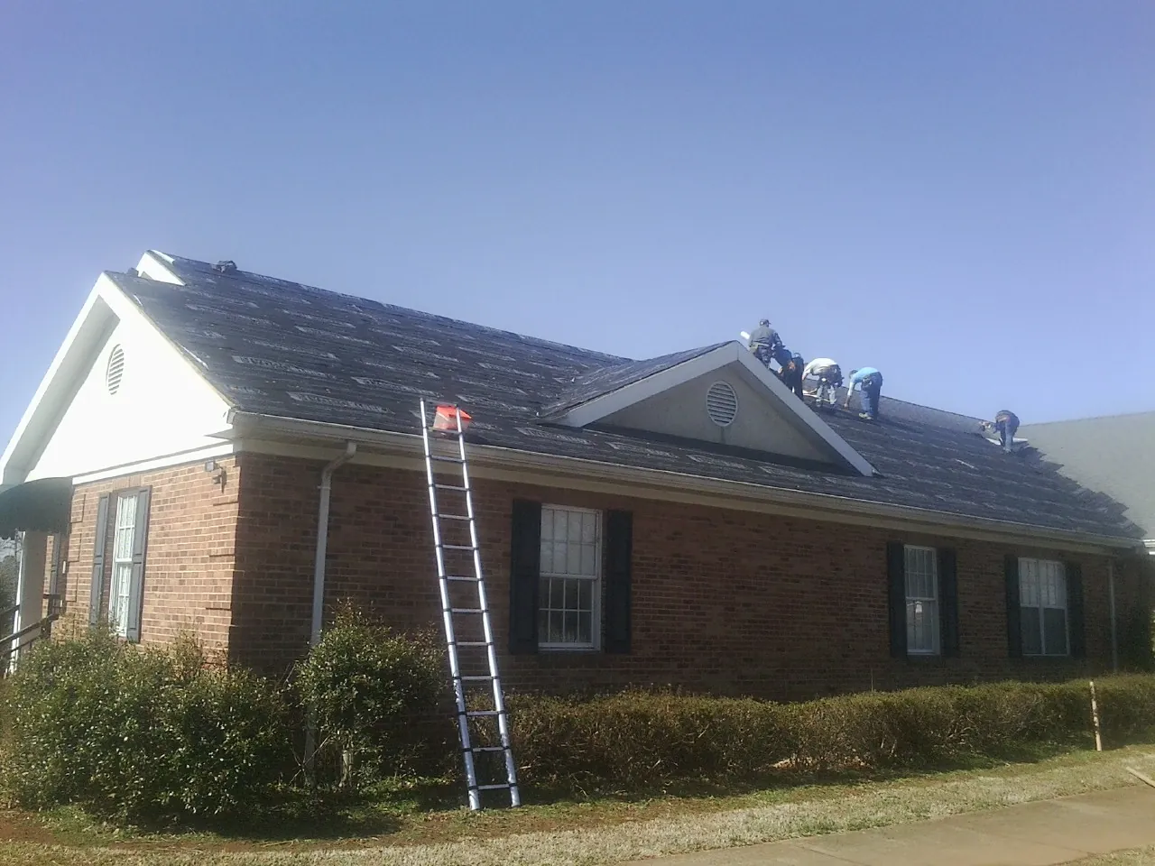 Crew tearing shingles off a brick ranch roof with a ladder set — Metal Roof Repair work by our crew in Joy