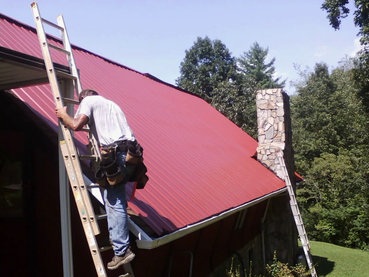 Roofer on a ladder beside a red metal roof and stone chimney — Soffit & Fascia Repair by Roofing Brothers in Joy